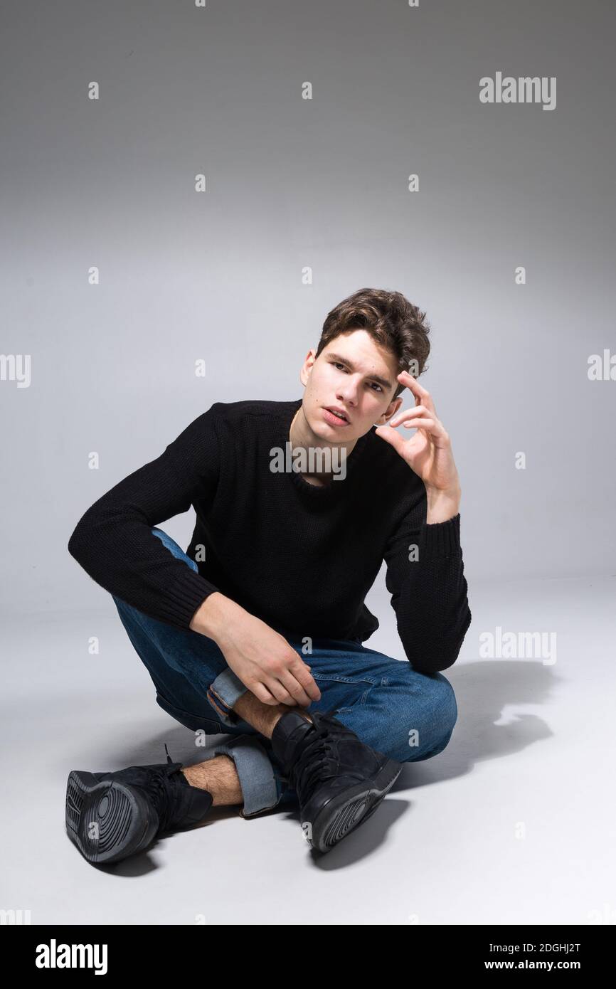 Elegante ragazzo in posa in studio seduto sul pavimento in pantaloni in denim e felpa nera. Ritratto di studente maschile pieno su sfondo bianco Foto Stock