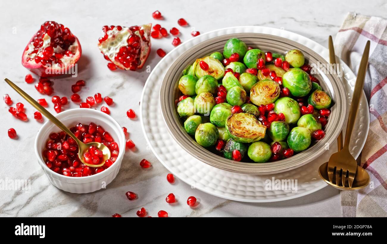 germogli di bruxelles arrostiti, salutare pasto vegetariano servito su un piatto bianco con posate dorate su fondo di marmo bianco con semi di melograno Foto Stock