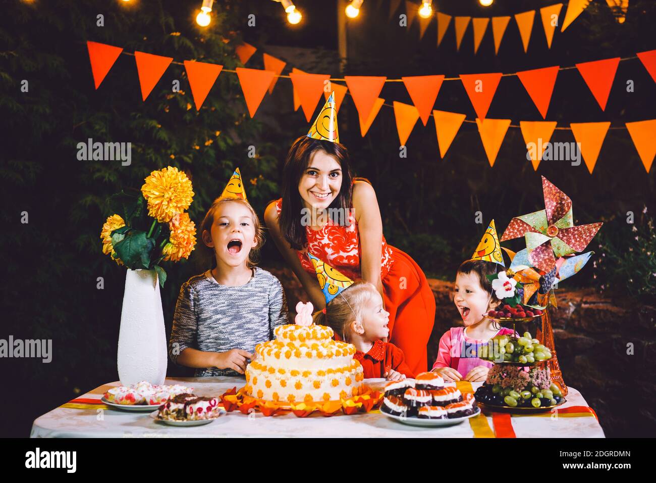 Una madre grande e tre bambini ad un tavolo di festa con dolci e una torta nel cortile decorato con luci e una ghirlanda Foto Stock
