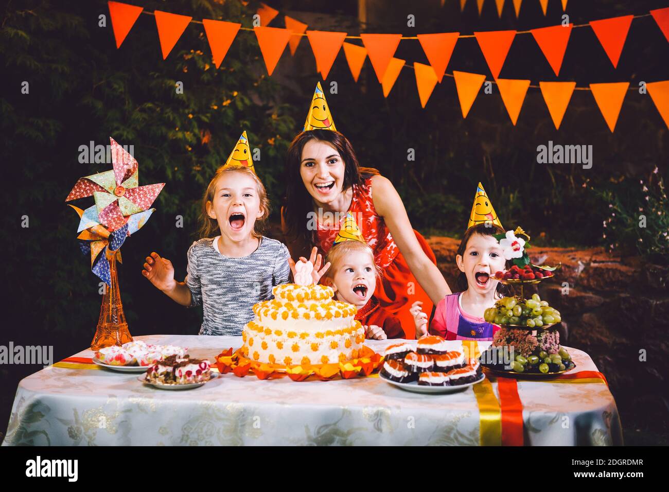 Una madre grande e tre bambini ad un tavolo di festa con dolci e una torta nel cortile decorato con luci e una ghirlanda Foto Stock