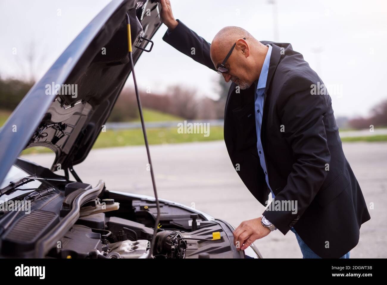 Professionista maturo elegante uomo d'affari preoccupato nella tuta sta guardando sotto la cappa dell'auto cercando di capire il problema. Foto Stock