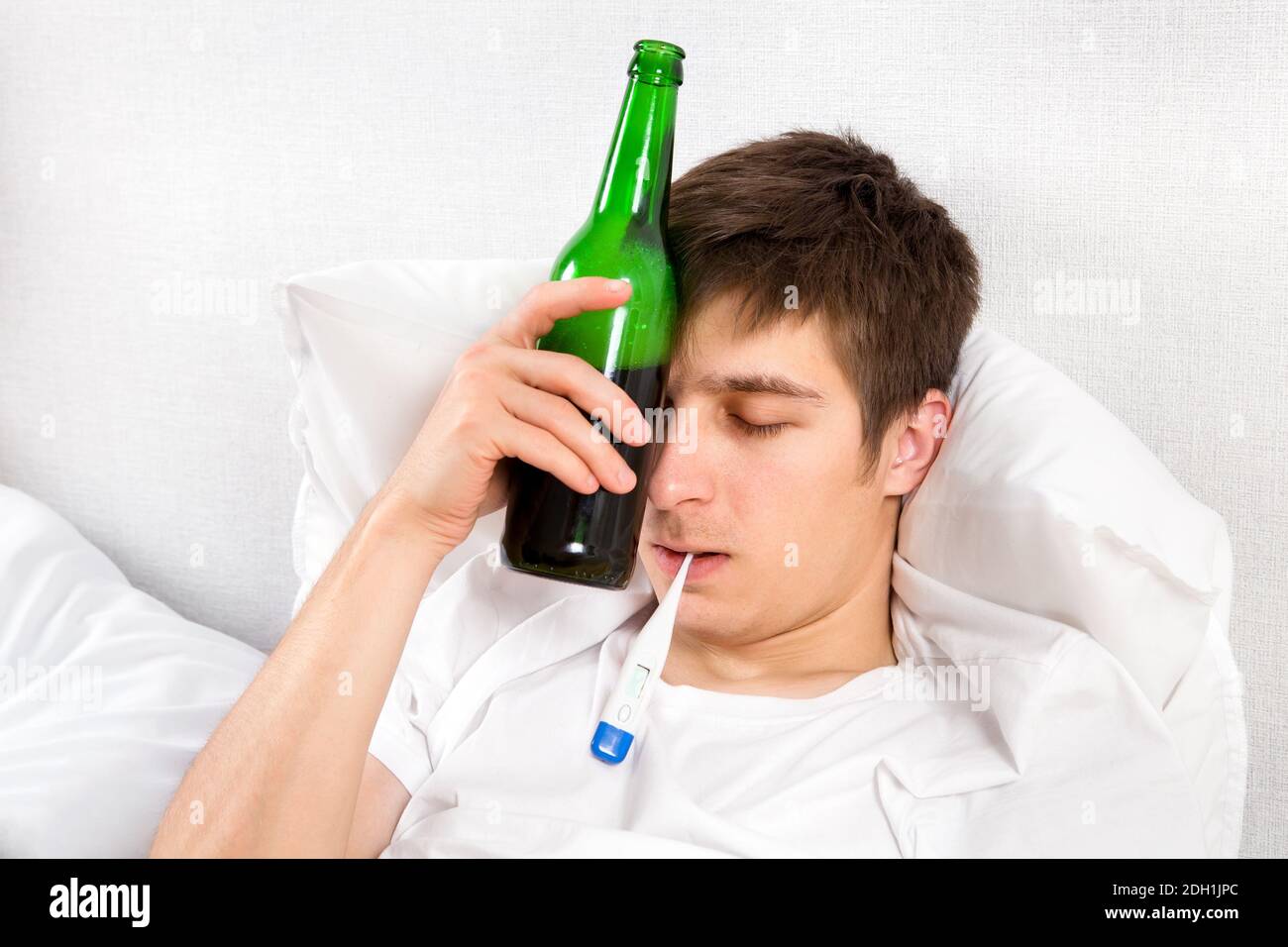 Giovane uomo con una bottiglia di birra e un termometro Il letto a casa Foto Stock