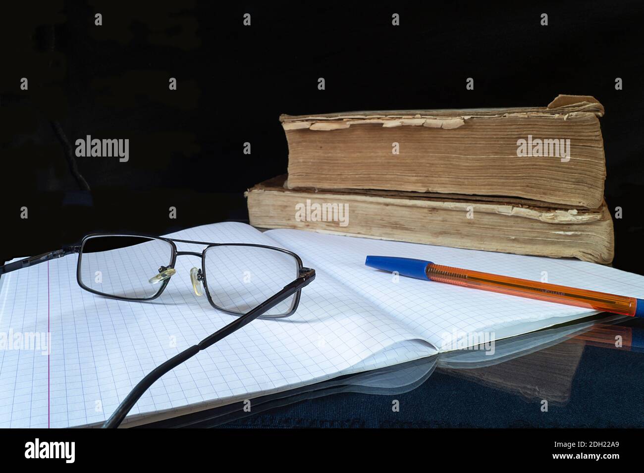 Gli occhiali, il blocco note e la maniglia da poggiare su un tavolo di vetro. I soggetti per imparare su sfondo nero. I libri antichi. Foto Stock