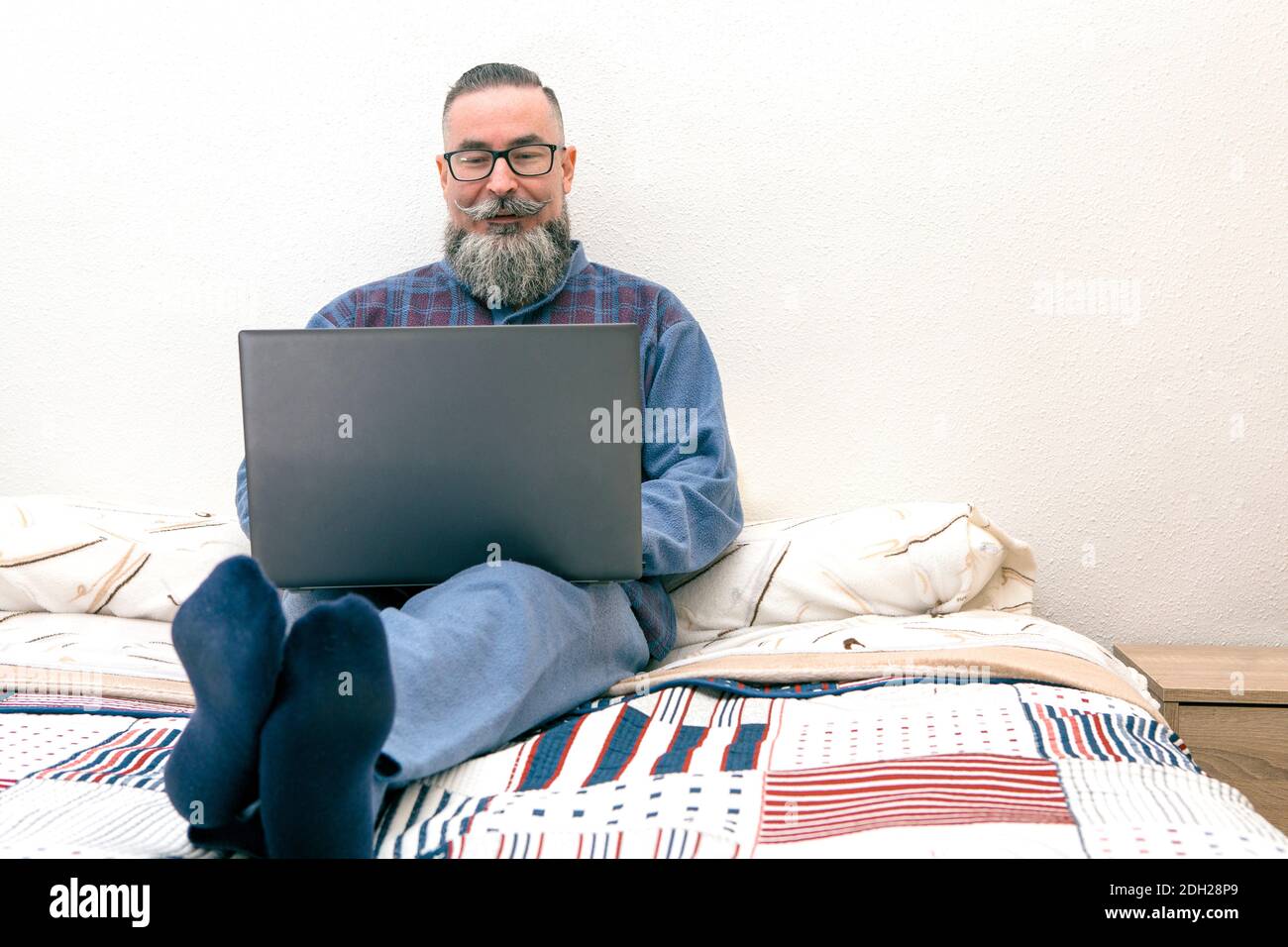 uomo con la barba grande seduto sul letto nel suo camera da letto con occhiali da pigiama e computer portatile Foto Stock