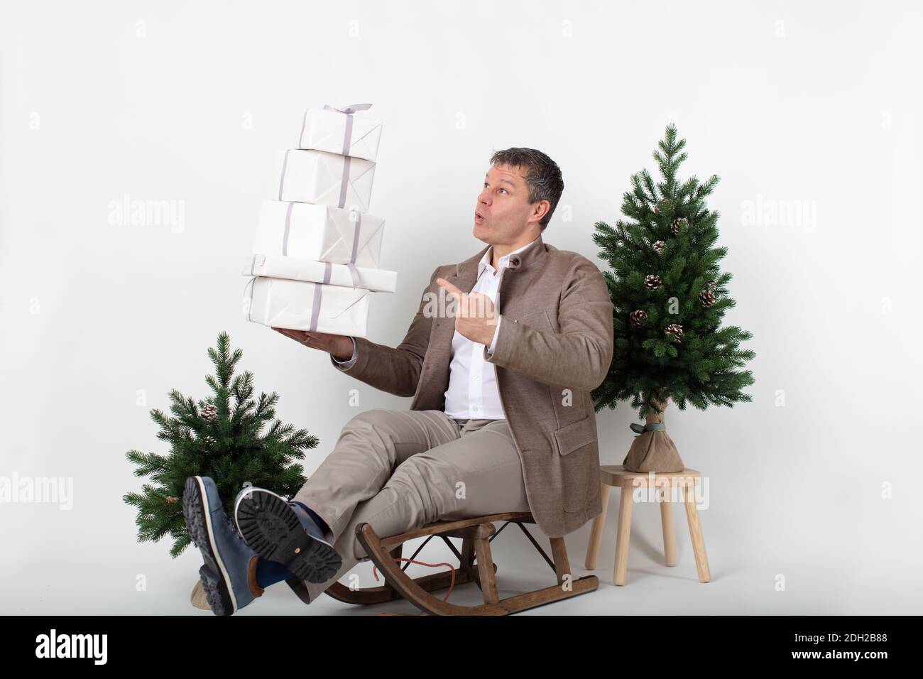 Ritratto di affari orizzontale a tema natalizio di un dirigente maschile elegante vestito e casual seduto su una slitta giocolante scatole regalo bianche avvolti, il tutto su uno sfondo bianco. Foto Stock