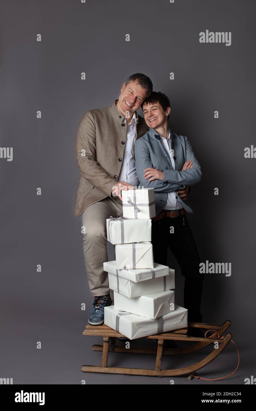 Ritratto di coppia di affari verticale a tema natalizio vestito casual elegante in pantaloni blu, bianco, beige e blazer su sfondo grigio. In primo piano una slitta di legno con un mucchio di scatole regalo bianche su di essa. Foto Stock