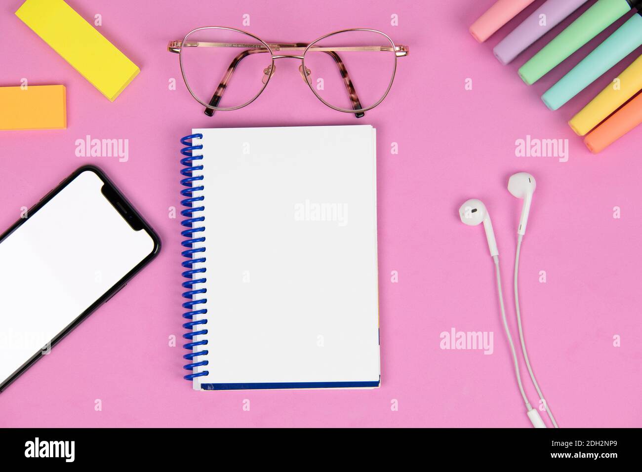 notebook vuoto su sfondo rosa con penna, telefono cellulare con schermo vuoto, note post-it colorate, cuffie, pennarelli colorati e occhiali Foto Stock