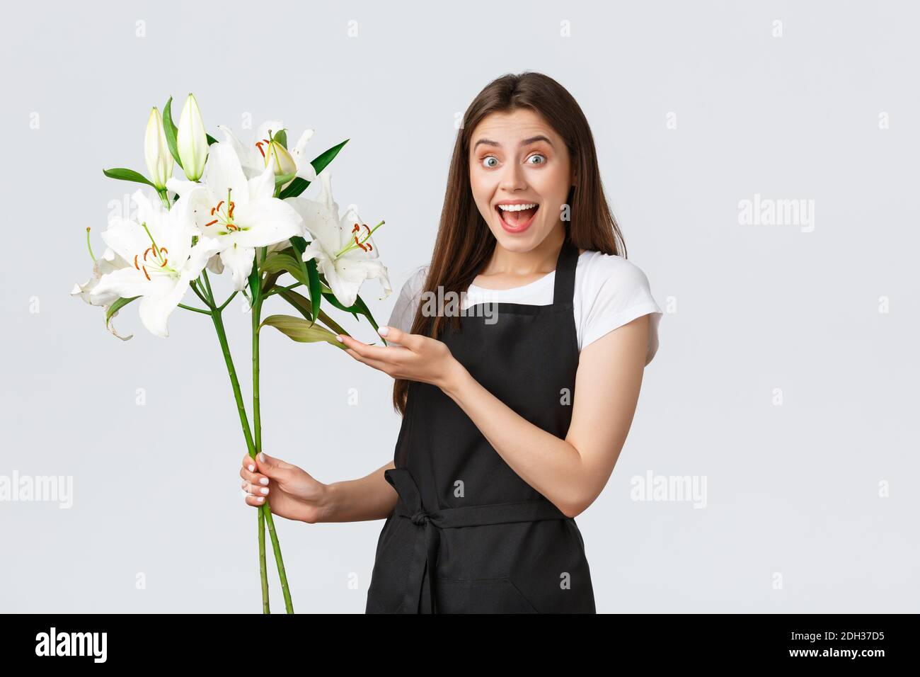 Concetto di shopping, dipendenti e piccole imprese. Entusiasta fiorista sorridente femminile in grembiule nero che guarda stupito come tenere bouq Foto Stock