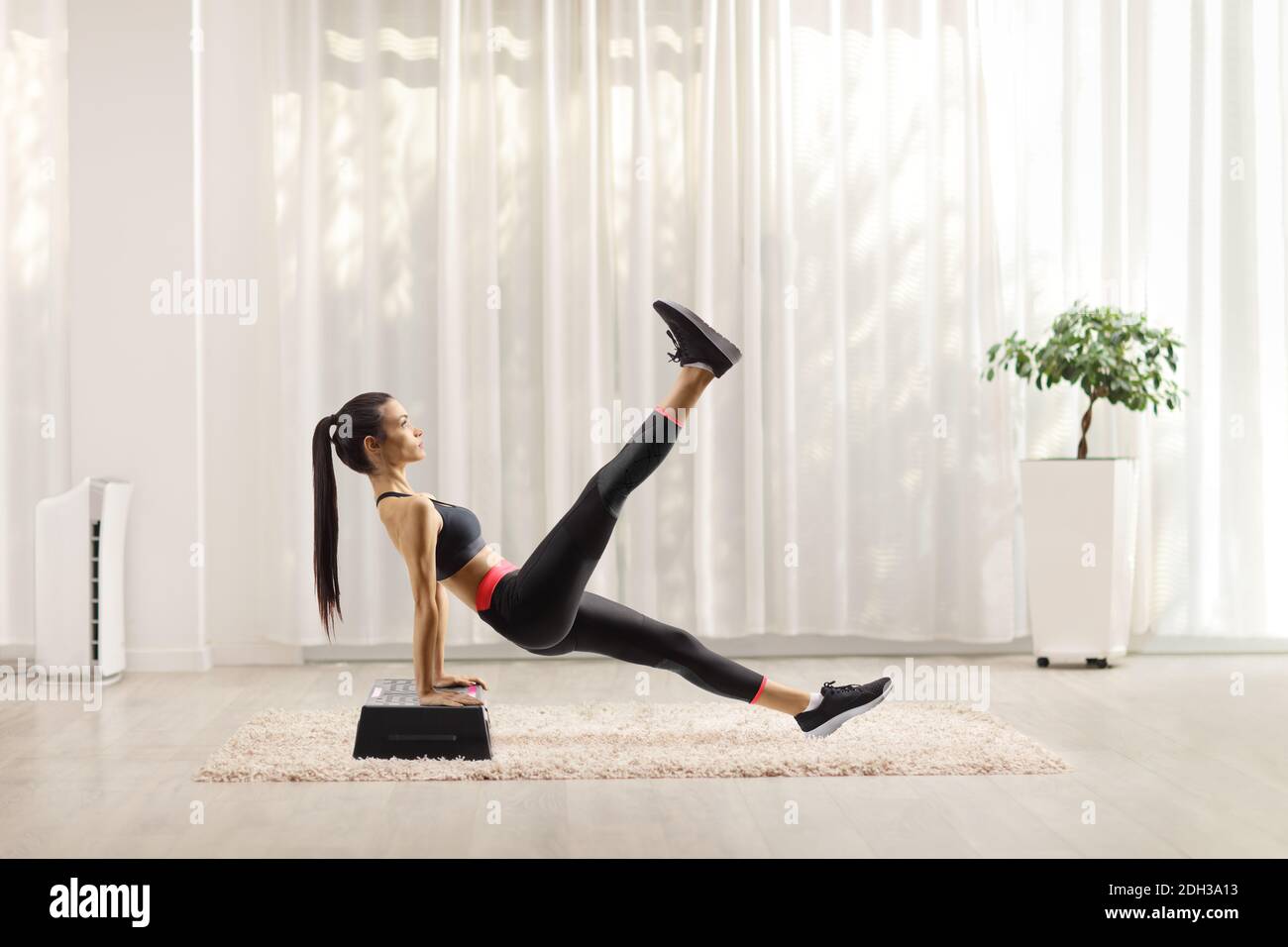 Foto di profilo a lunghezza intera di una giovane donna che si esercita su una piattaforma aerobica di punto e sollevando una gamba a casa Foto Stock