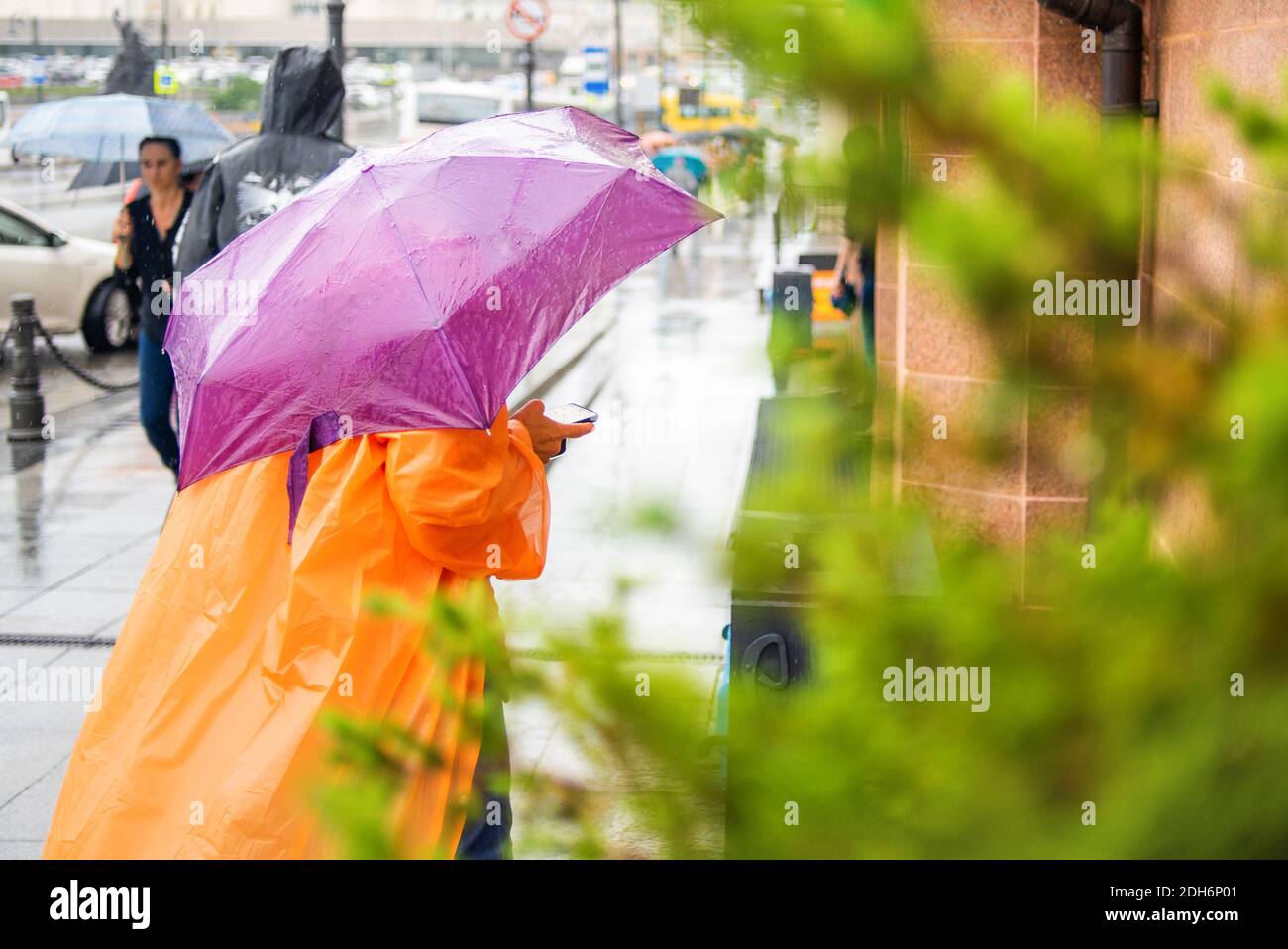 Donna in un impermeabile arancione con un ombrello viola tiene un telefono in mano. Paesaggio urbano in tempo piovoso. Giorno piovoso. Maltempo. Foto Stock