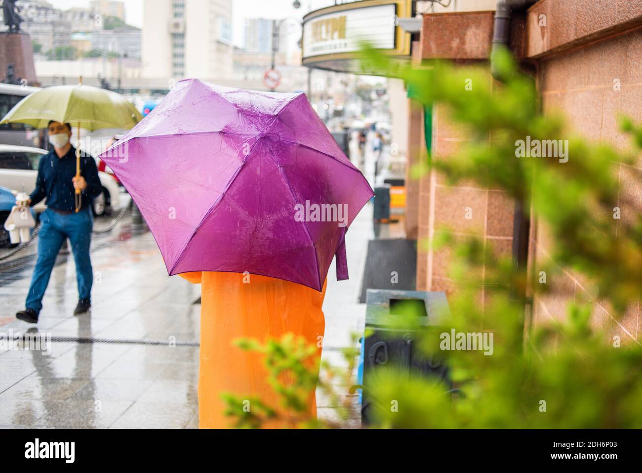 Donna in un impermeabile arancione con ombrello viola in tempo piovoso sullo sfondo della città. Paesaggio urbano in cattive condizioni atmosferiche. Giorno piovoso. Stile cittadino. Foto Stock
