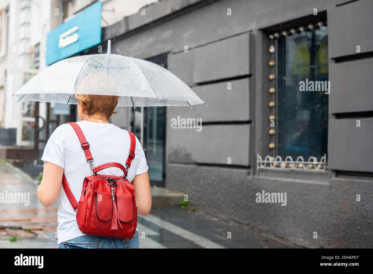 Donna con un ombrello trasparente cammina per la città in tempo piovoso. Ombrello con gocce di pioggia. Scene della città nel maltempo piovoso. Foto Stock