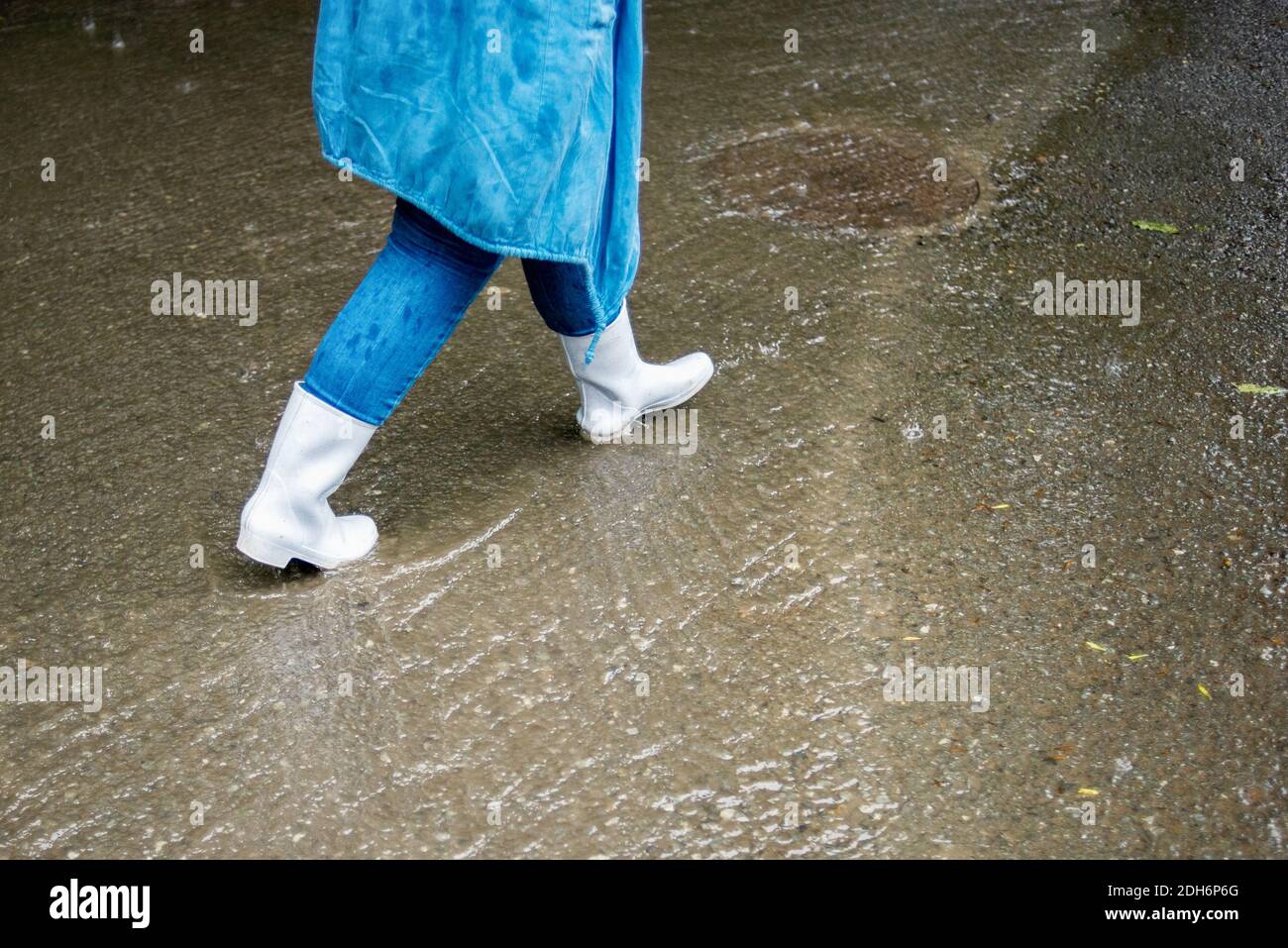 Una donna in stivali di gomma bianca cammina per la strada sotto la pioggia. L'acqua scorre lungo la strada. Scene di strada sotto la pioggia. Tempo di caduta piovoso. Foto Stock