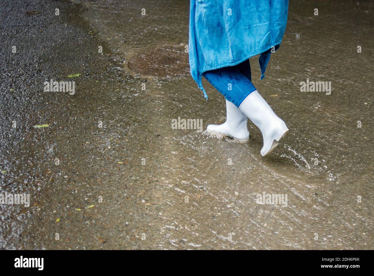 Una donna in stivali di gomma bianca cammina per la strada sotto la pioggia. L'acqua scorre lungo la strada. Scene di strada sotto la pioggia. Tempo di autunno piovoso. Foto Stock