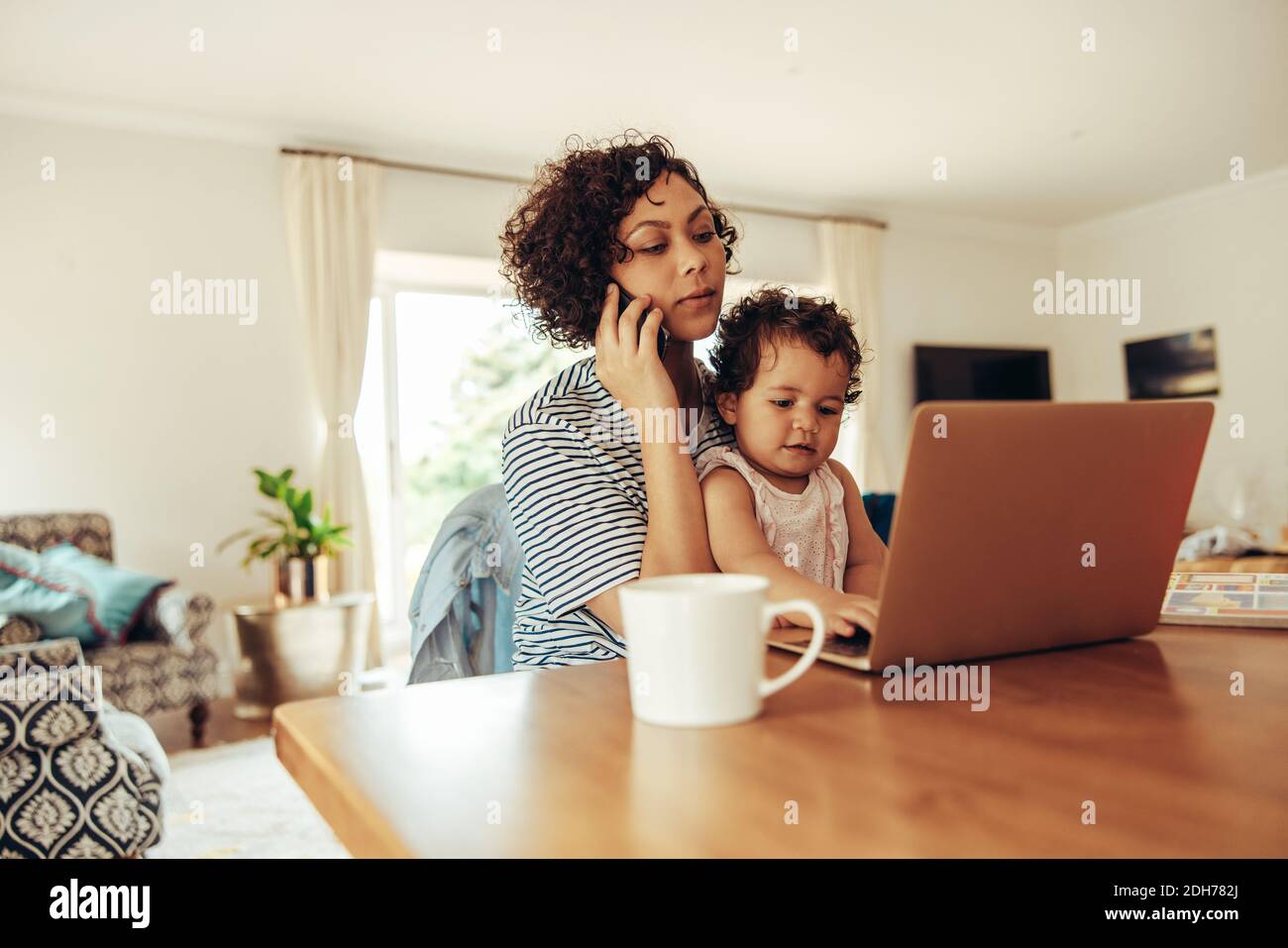 Donna freelance che usa il cellulare e il laptop con il suo bambino seduto in grembo a casa. Madre che lavora a casa con il bambino. Foto Stock