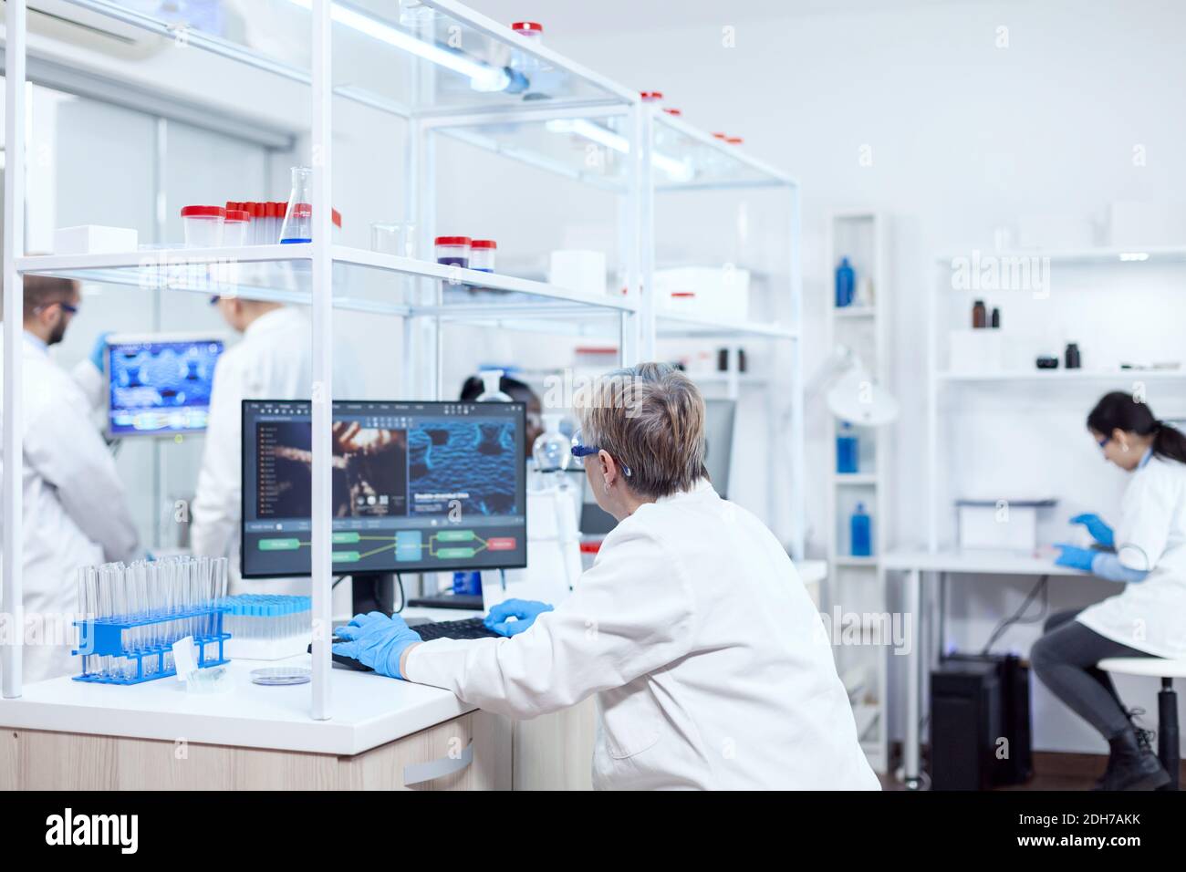 Un chimico medico anziano anziano che lavora su un potente computer in una struttura moderna. Scienziato senior in laboratorio farmaceutico che fa ricerche genetiche indossando camice da laboratorio con squadra in background. Foto Stock