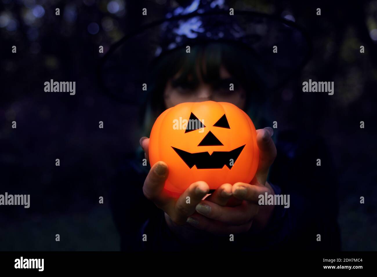 Zucca di Halloween tenuta da mani magiche a mezzanotte. Una zucca incandescente con gli occhi e la bocca, con un sorriso malvagio e una strega sfocata sullo sfondo. Foto Stock
