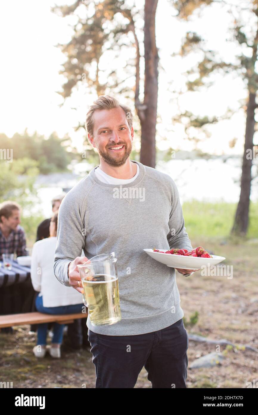 Ritratto di felice uomo che tiene la brocca di birra e fragole con amici seduti al tavolo da picnic in background Foto Stock