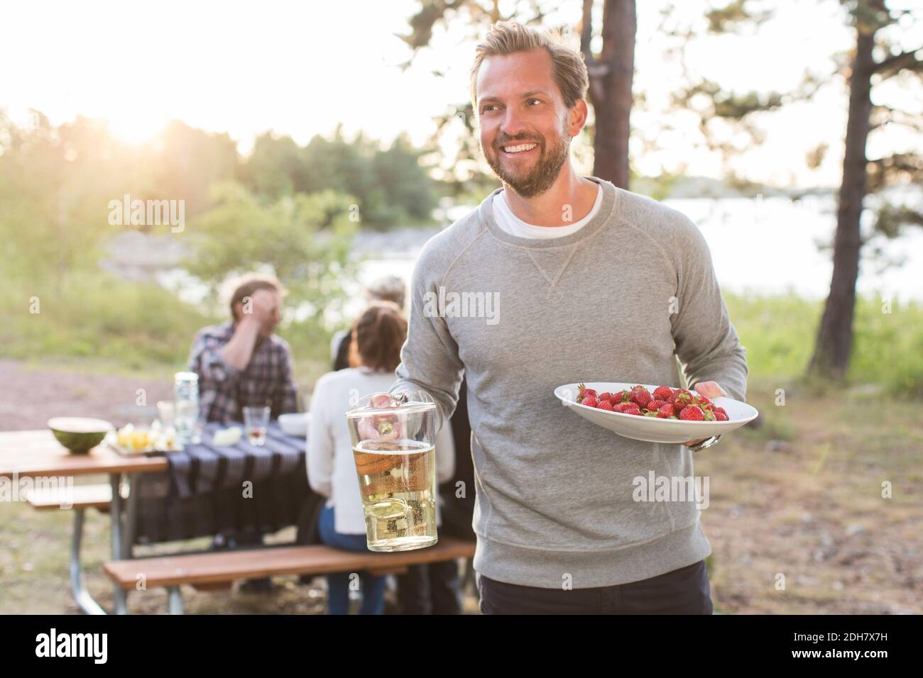 Felice uomo che tiene la brocca di birra e fragole con gli amici seduti al tavolo da picnic in background Foto Stock