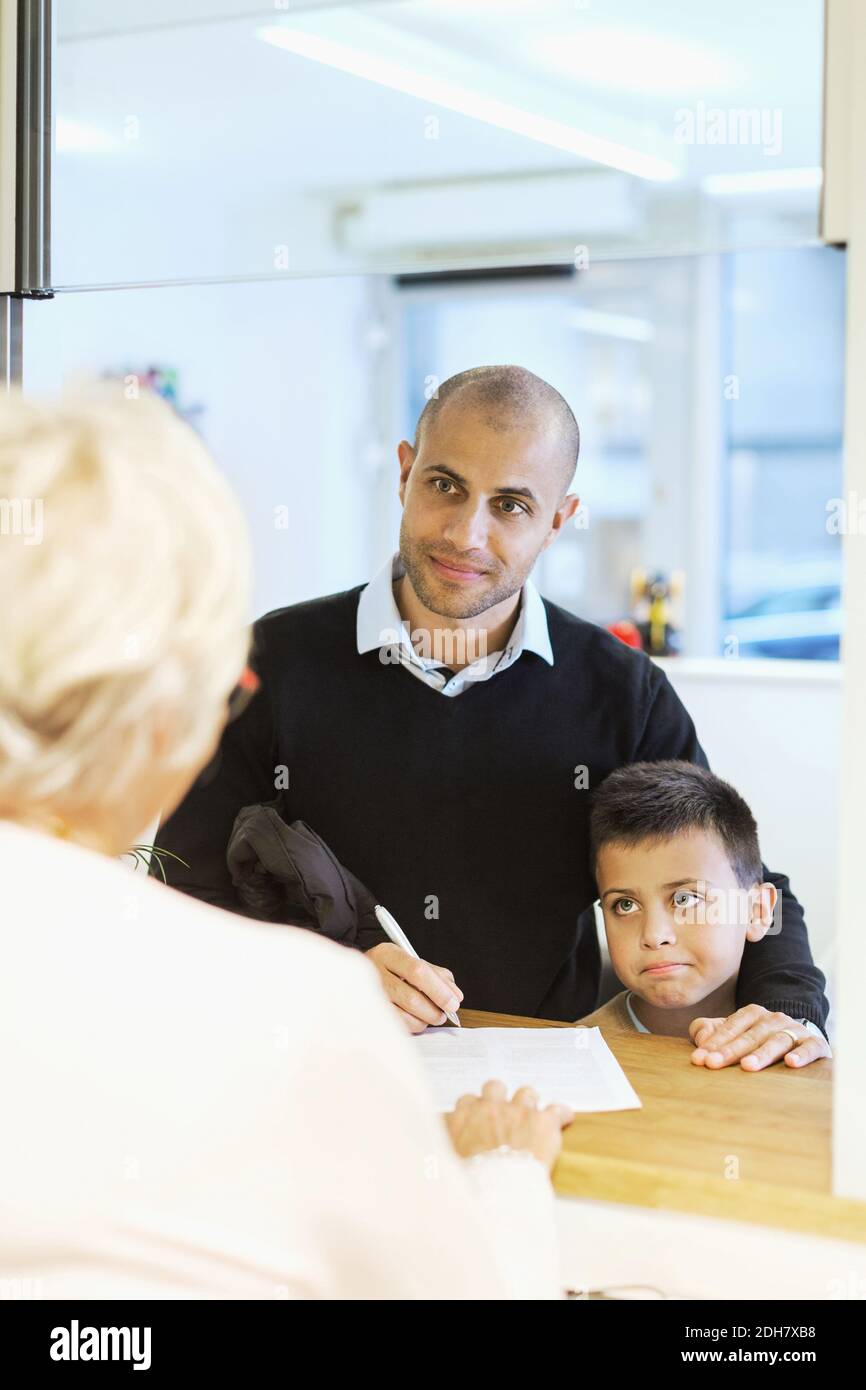 Padre e figlio guardano l'addetto alla reception mentre riempiono i moduli ambulatorio ortopedico Foto Stock