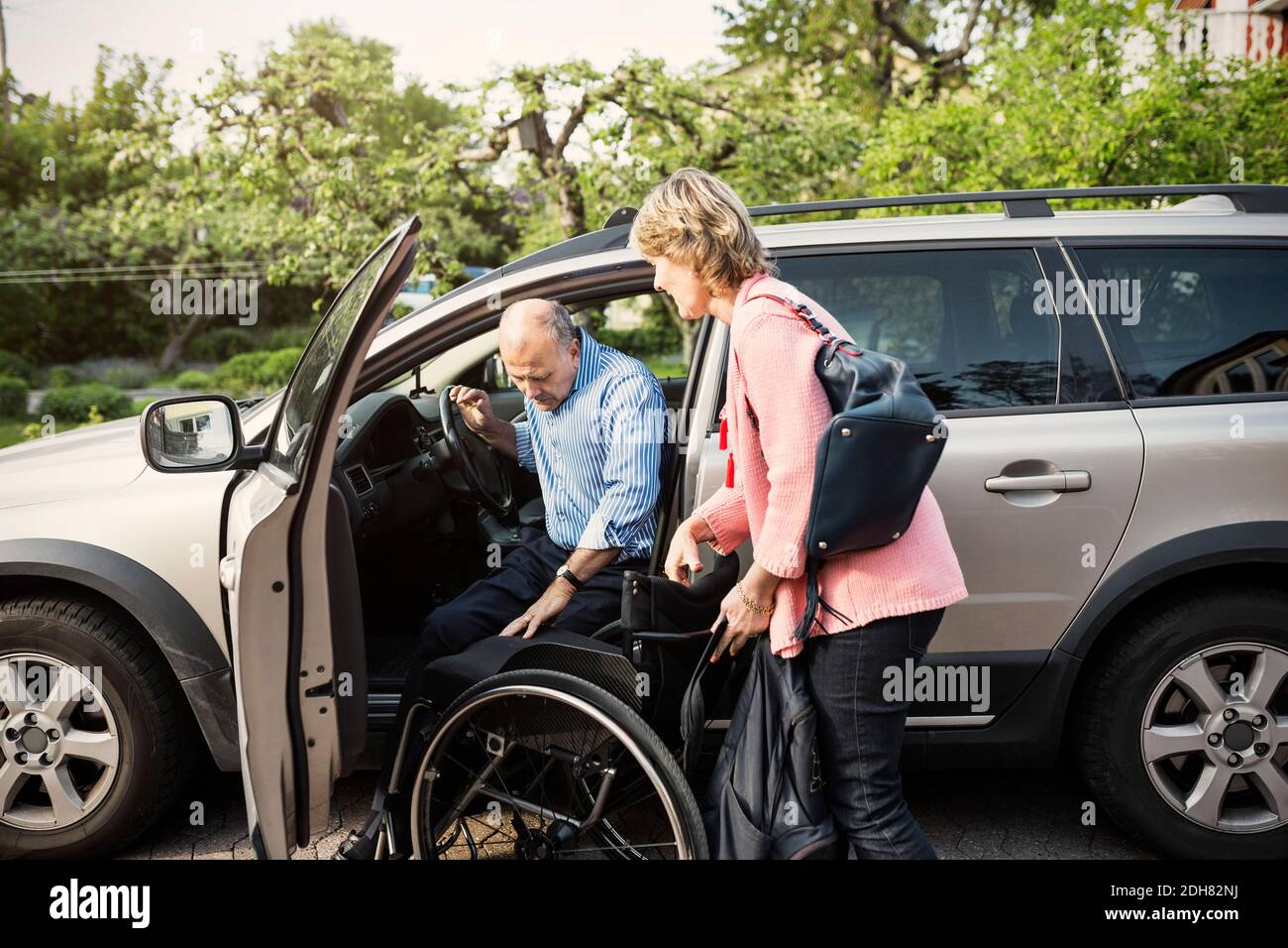 Uomo disabile che sbarca dall'auto su sedia a rotelle tenuto da donna Foto Stock