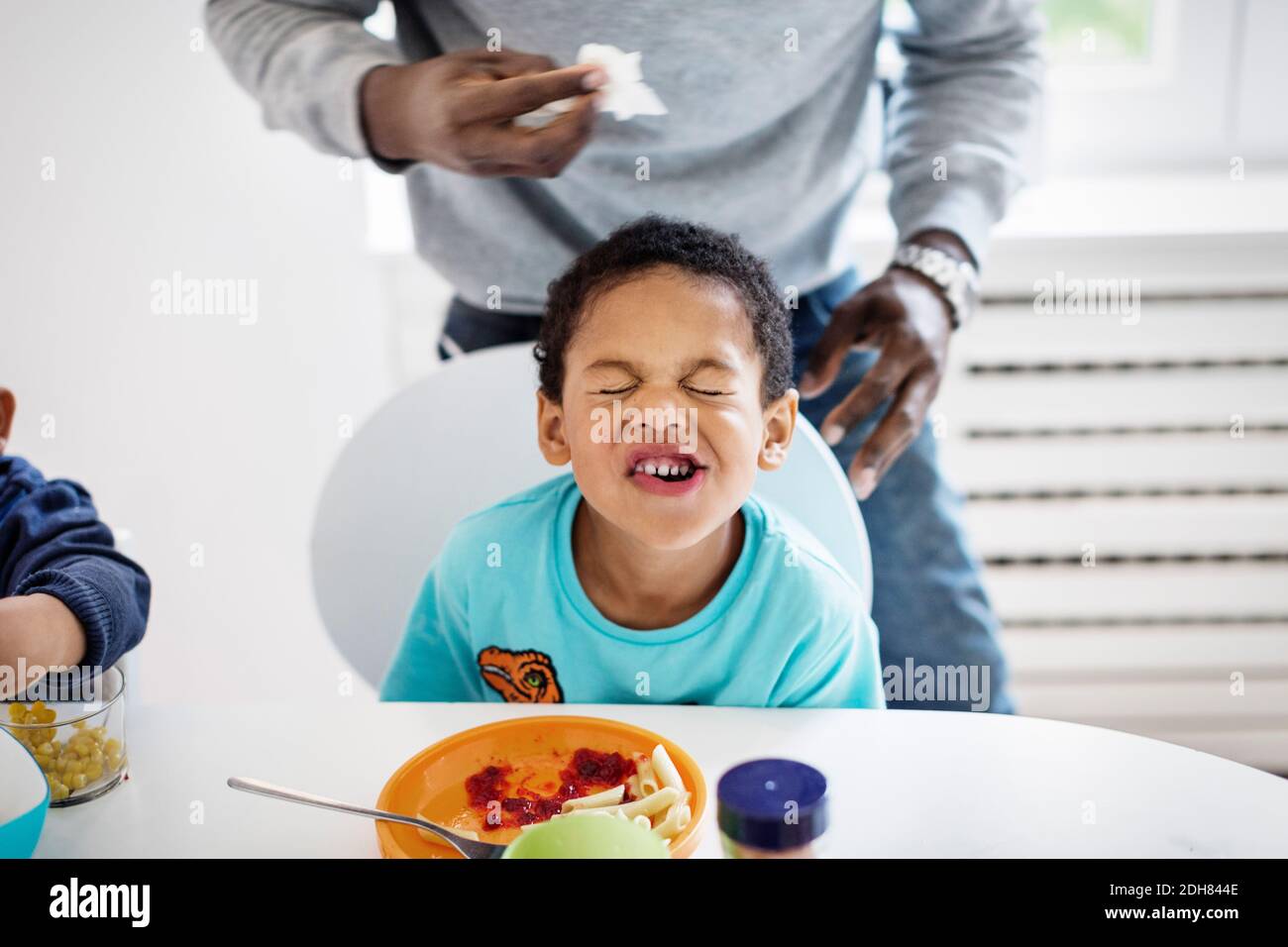 Ragazzo che fa la faccia mentre ha il cibo a tavola con il padre in piedi sullo sfondo Foto Stock