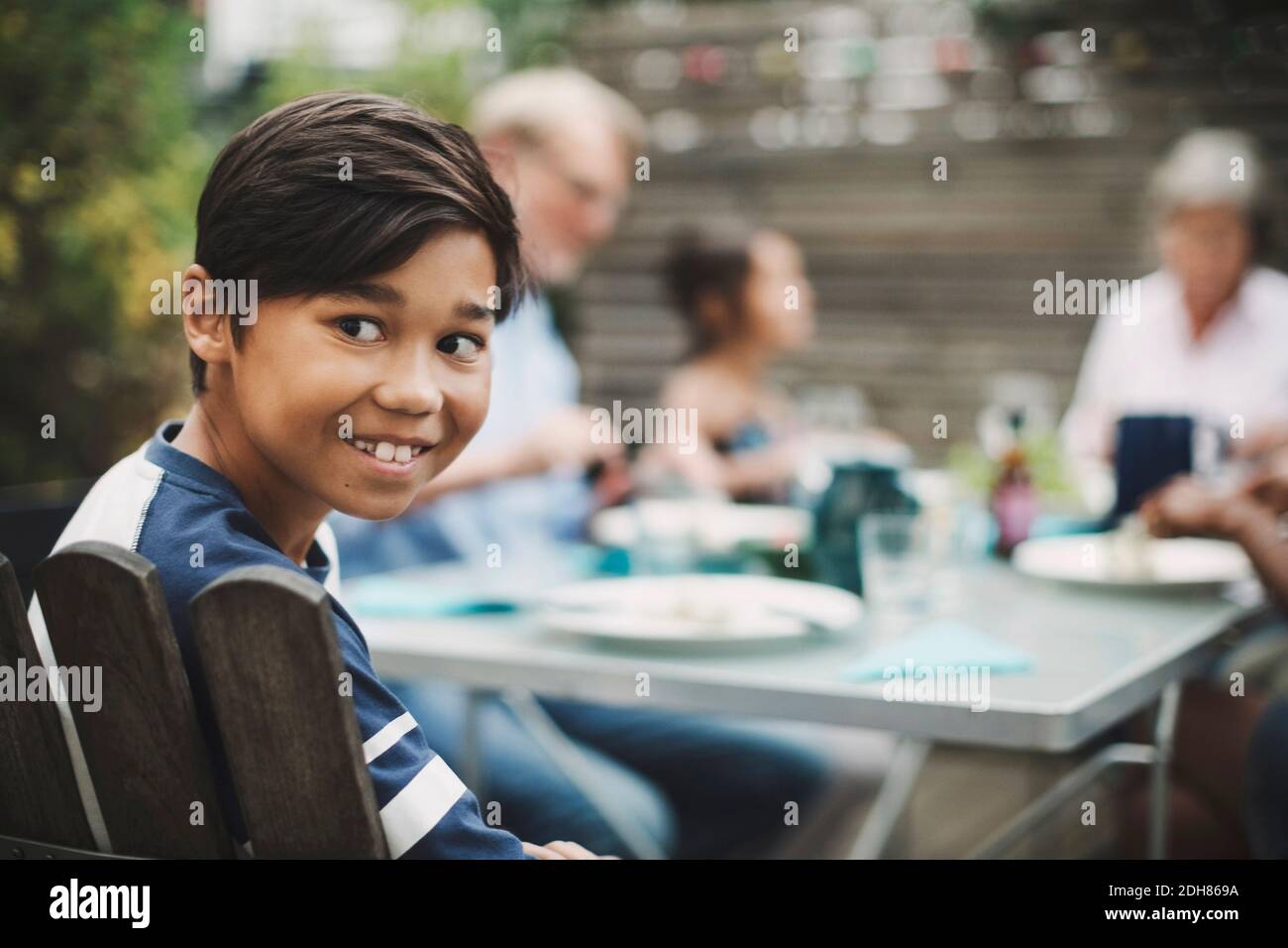 Ragazzo felice che guarda via mentre si siede con la famiglia a pranzo tavolo in cortile Foto Stock