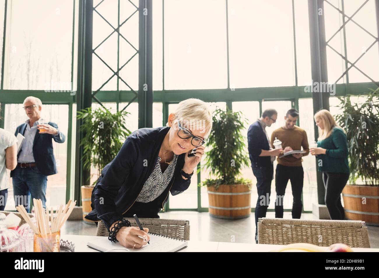 Donna d'affari matura su chiamata mentre scrive in blocco note con i colleghi in background in ufficio Foto Stock