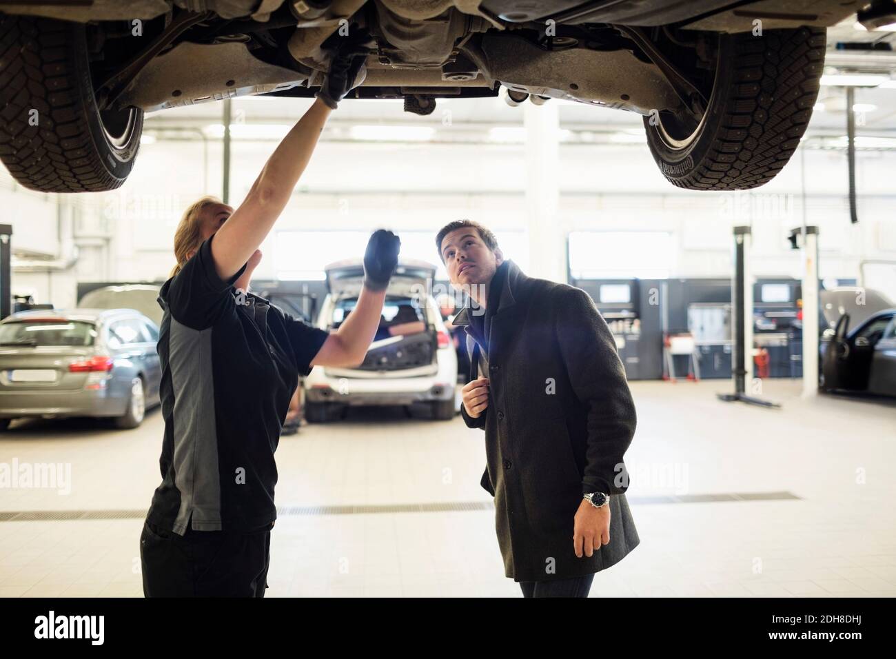 Meccanico femminile che discute con il cliente sotto l'auto al negozio di riparazione Foto Stock