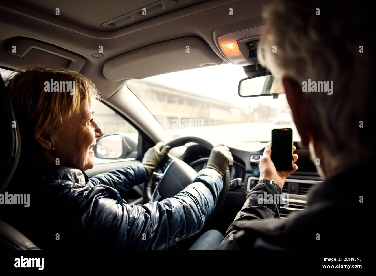 Donna anziana sorridente che guida l'auto mentre si siede da un uomo che prende selfie tramite smartphone Foto Stock