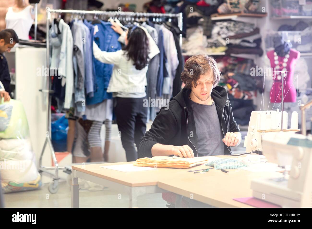 Uomo che lavora su macchina da cucire mentre volontario cerca i vestiti in rack in officina Foto Stock