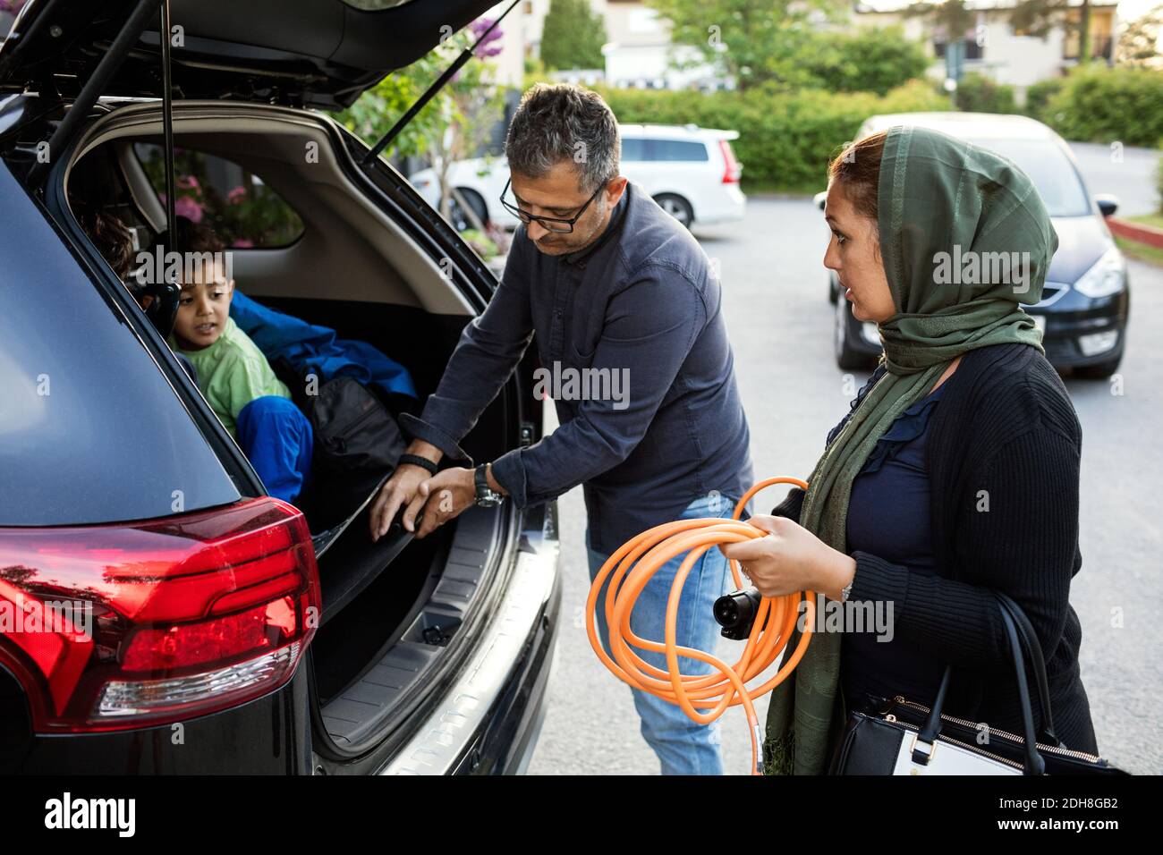 Madre che tiene il tubo mentre il padre che tiene il caricatore elettrico da figlio in auto Foto Stock