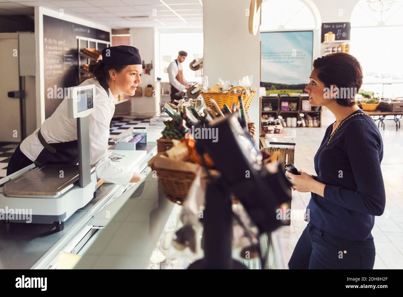 Vista laterale della donna che parla con il venditore al banco a. negozio di alimentari Foto Stock