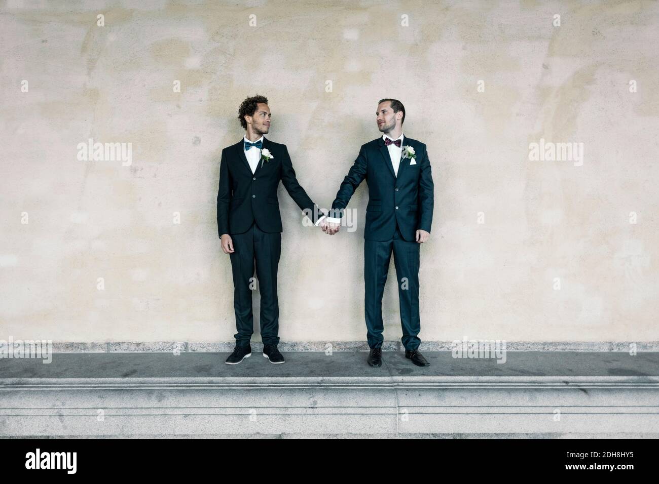 Coppia gay newlywed in piedi mentre tiene le mani sulla panchina contro parete Foto Stock