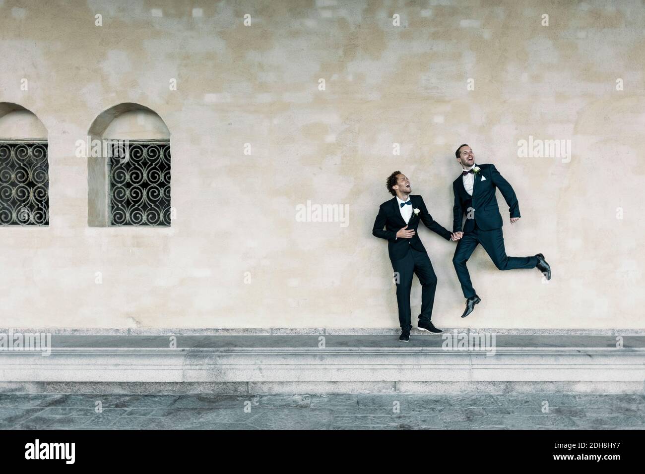 Uomo allegro che salta mentre tiene le mani del compagno gay sopra panca contro parete Foto Stock