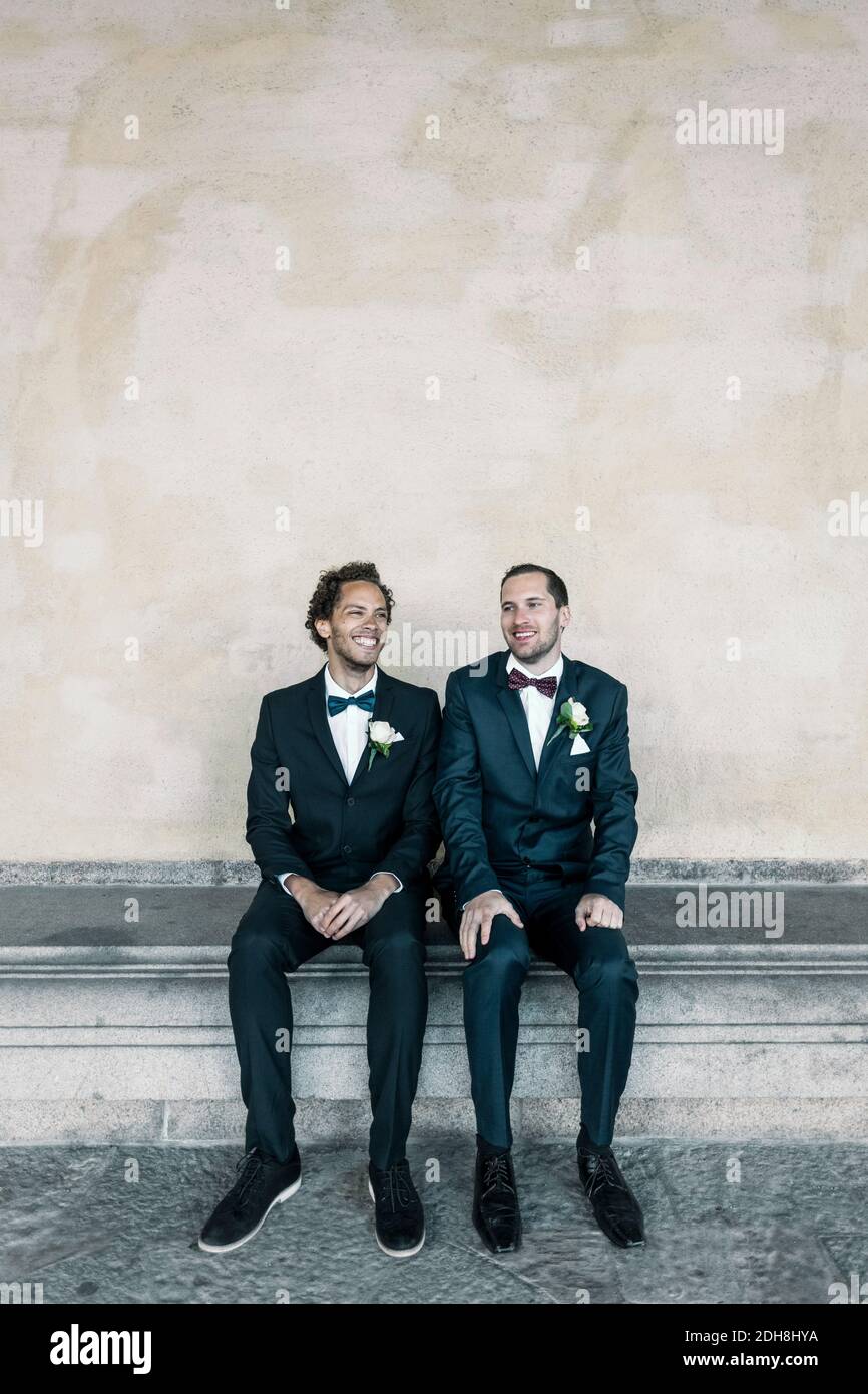 Felice coppia gay newlywed seduta su panchina contro muro Foto Stock