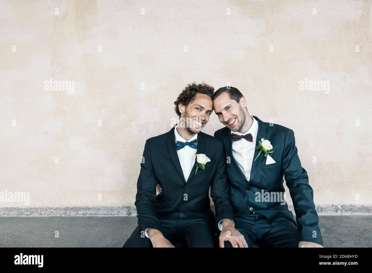 Ritratto di una coppia gay appena sposata seduta su una panchina contro un muro Foto Stock