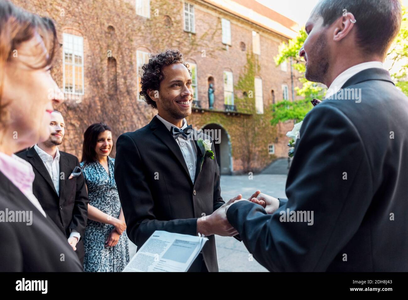 Sacerdote e amici che guardano una coppia gay che tiene le mani durante cerimonia nuziale Foto Stock