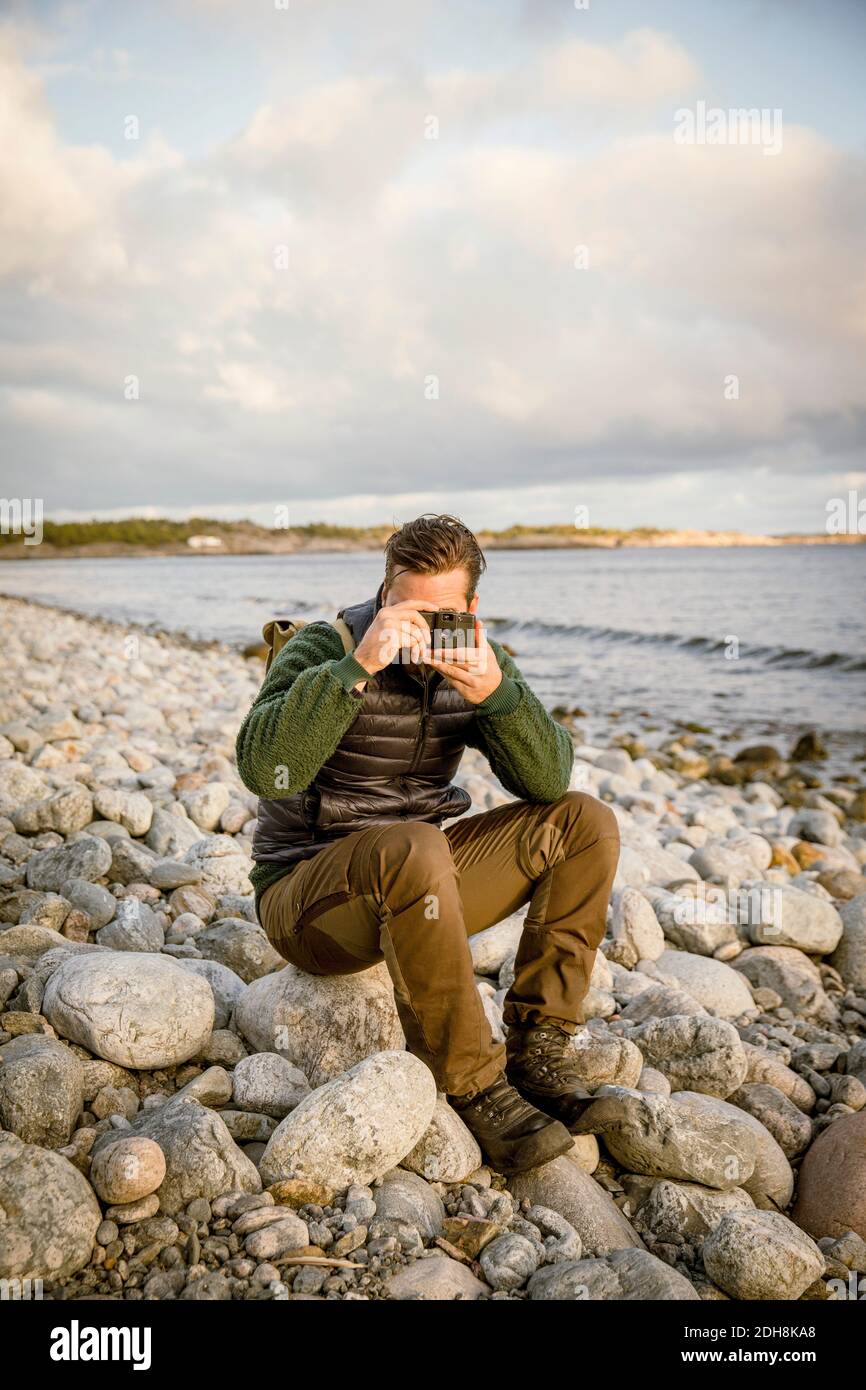 Uomo che fotografa dalla macchina fotografica mentre si siede sulla roccia in spiaggia contro il cielo Foto Stock