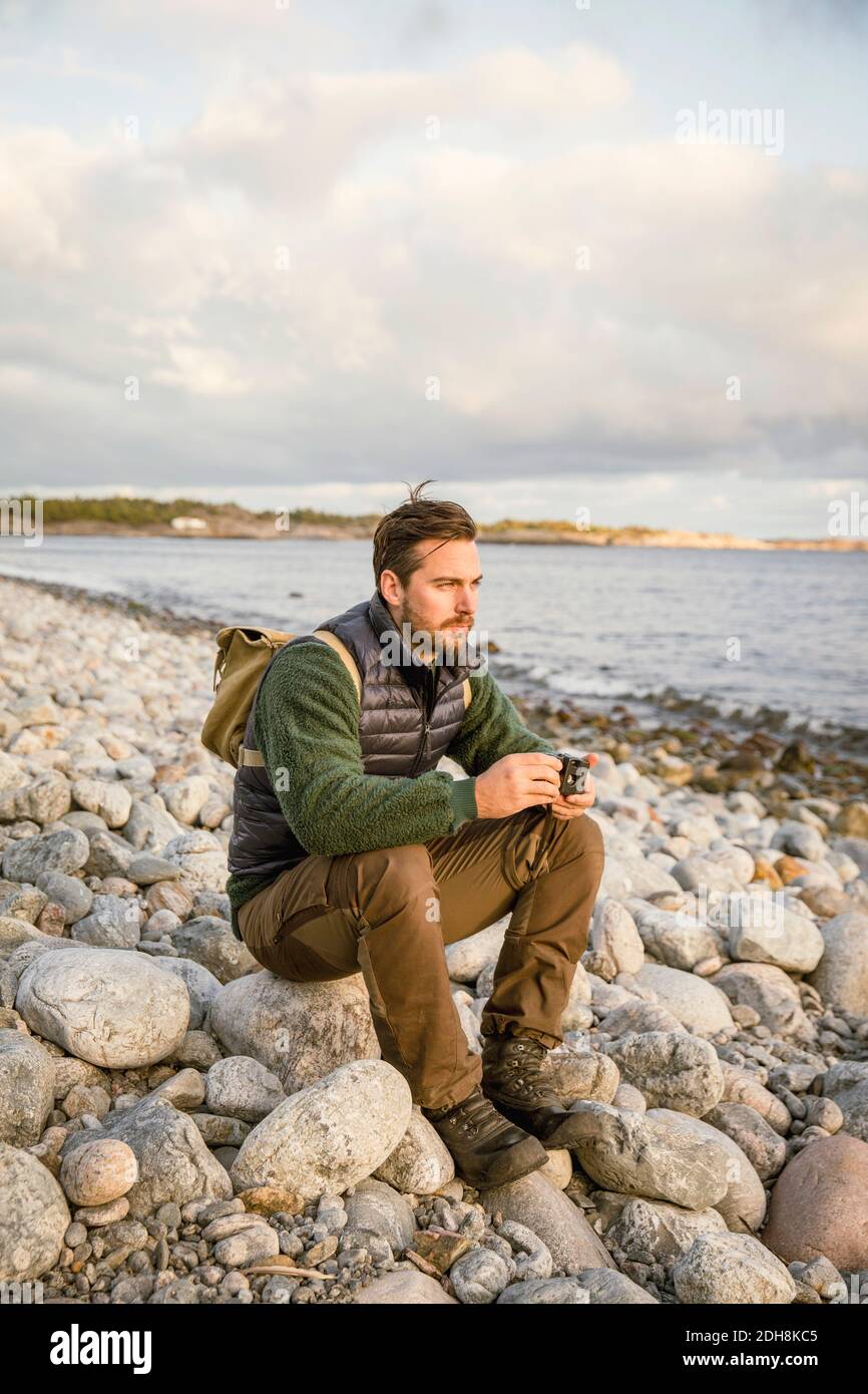 Uomo seduto sulla roccia con macchina fotografica sulla spiaggia contro il cielo Foto Stock