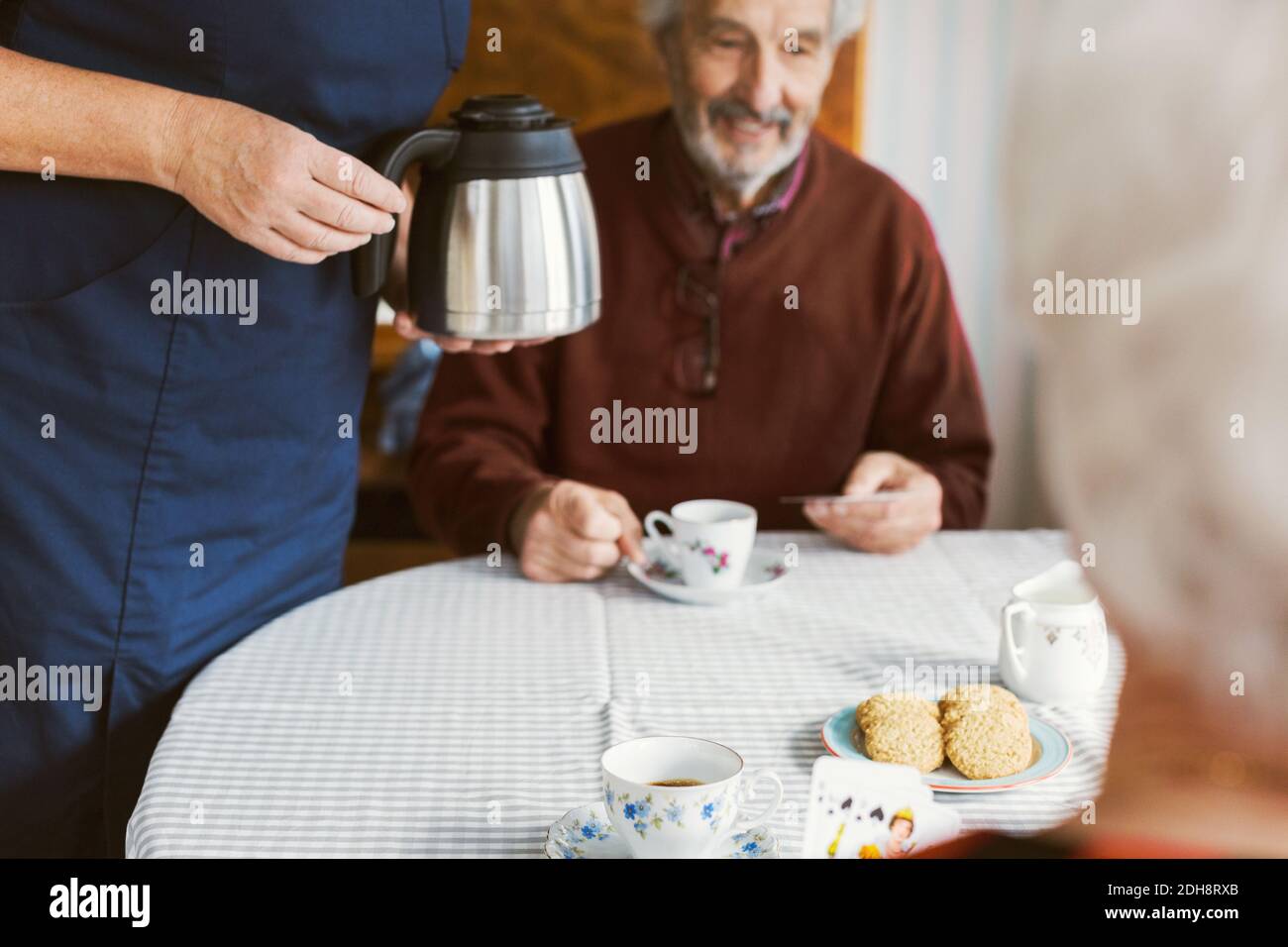 A metà del curatore che tiene la caffettiera mentre la coppia anziana gioca schede in casa di cura Foto Stock