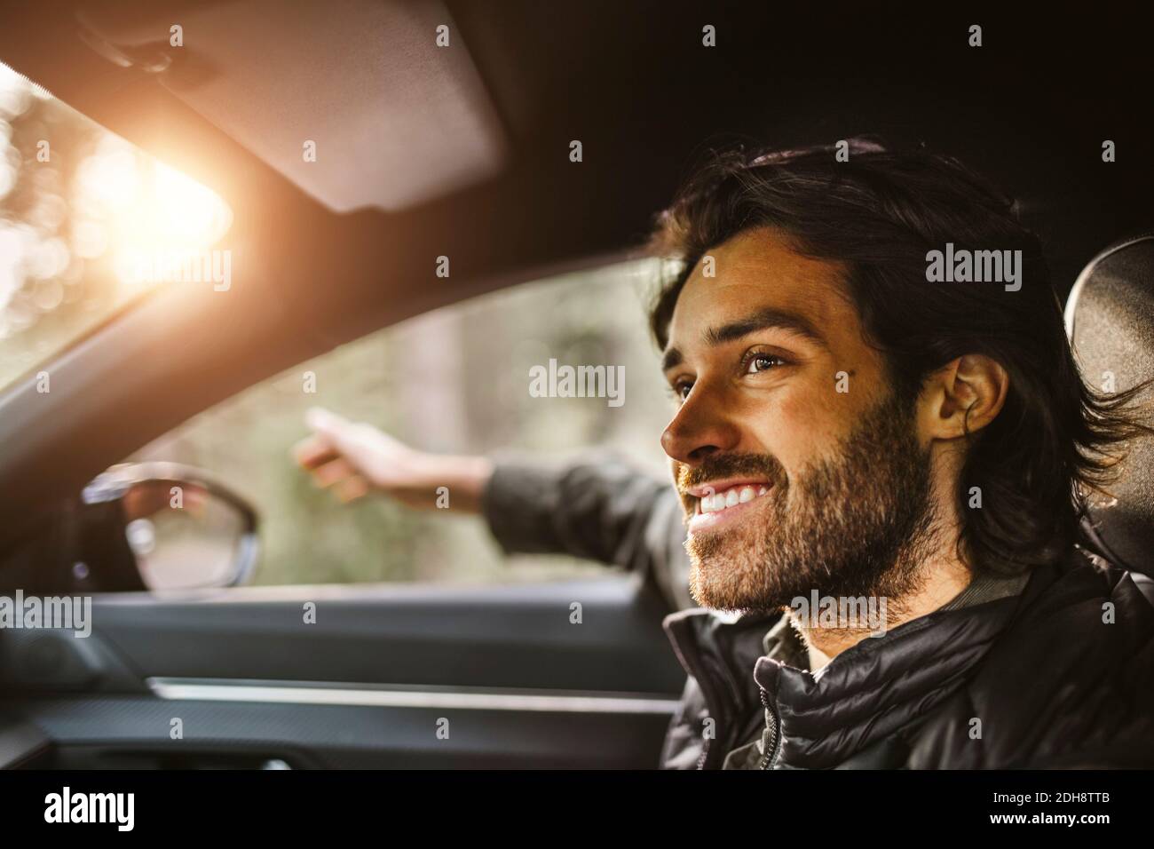 Uomo sorridente con mano fuori dalla finestra dell'auto guardando via durante viaggio su strada Foto Stock
