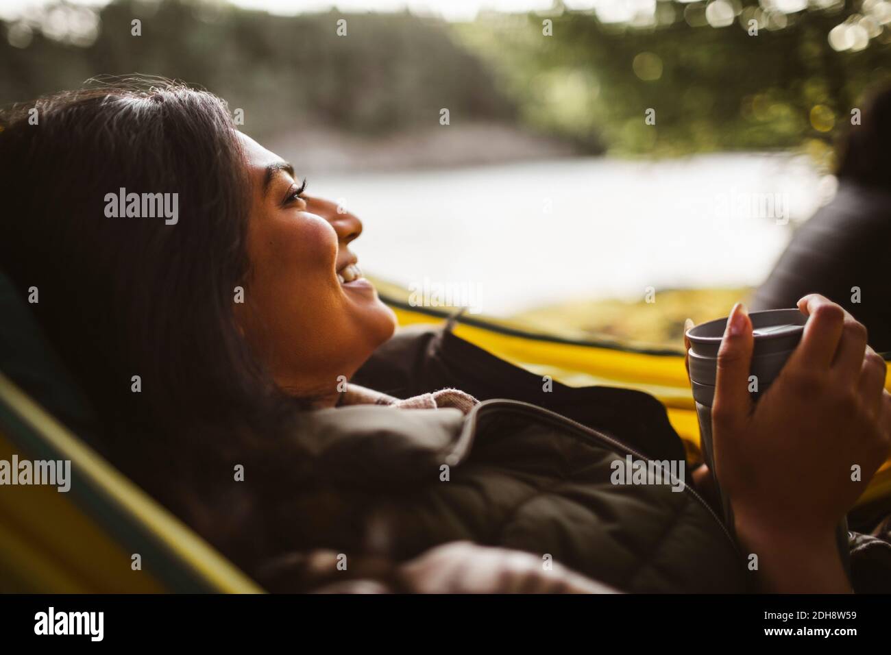 Donna sorridente con caffè che guarda via sull'amaca nella foresta durante le vacanze Foto Stock