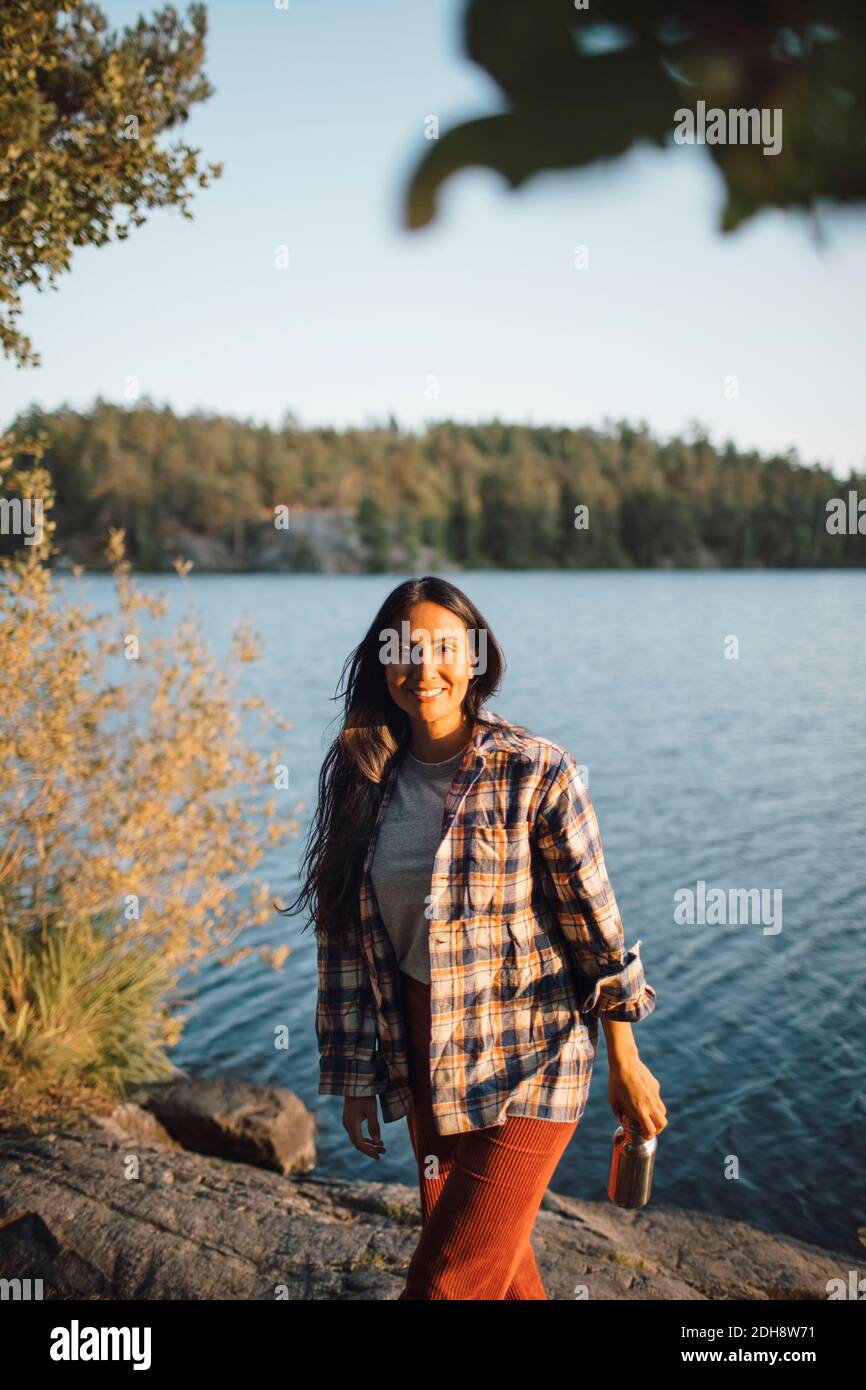 Ritratto di donna sorridente con bottiglia contro il lago nella foresta Foto Stock