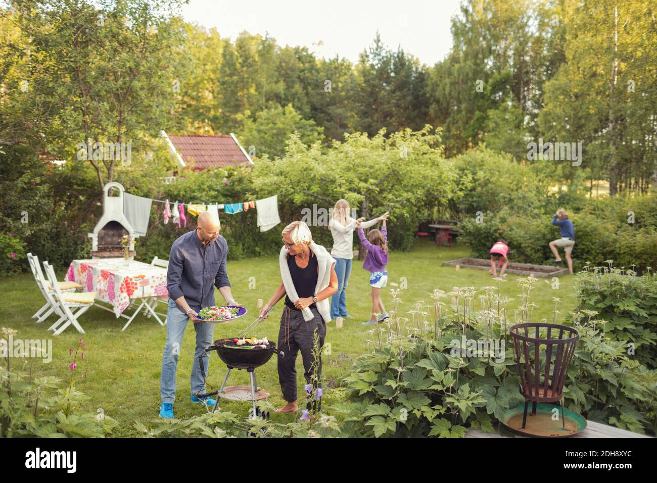 Uomo e donna cucinano verdure alla griglia con i bambini godetevi una festa in giardino Foto Stock