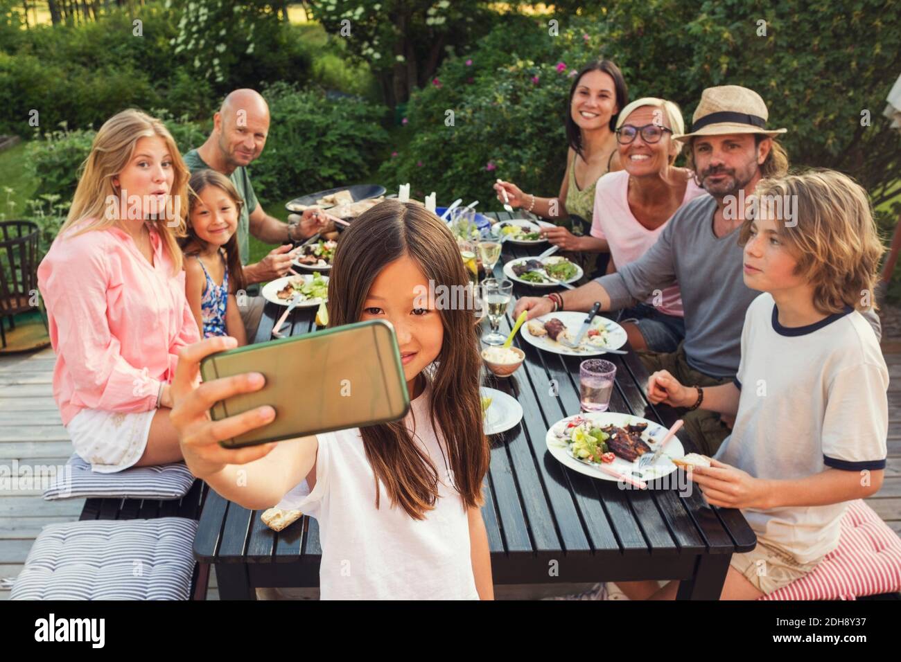 Famiglia felice e amici che cercano mentre la ragazza prende selfie da telefono cellulare nel cortile posteriore durante la festa in giardino Foto Stock