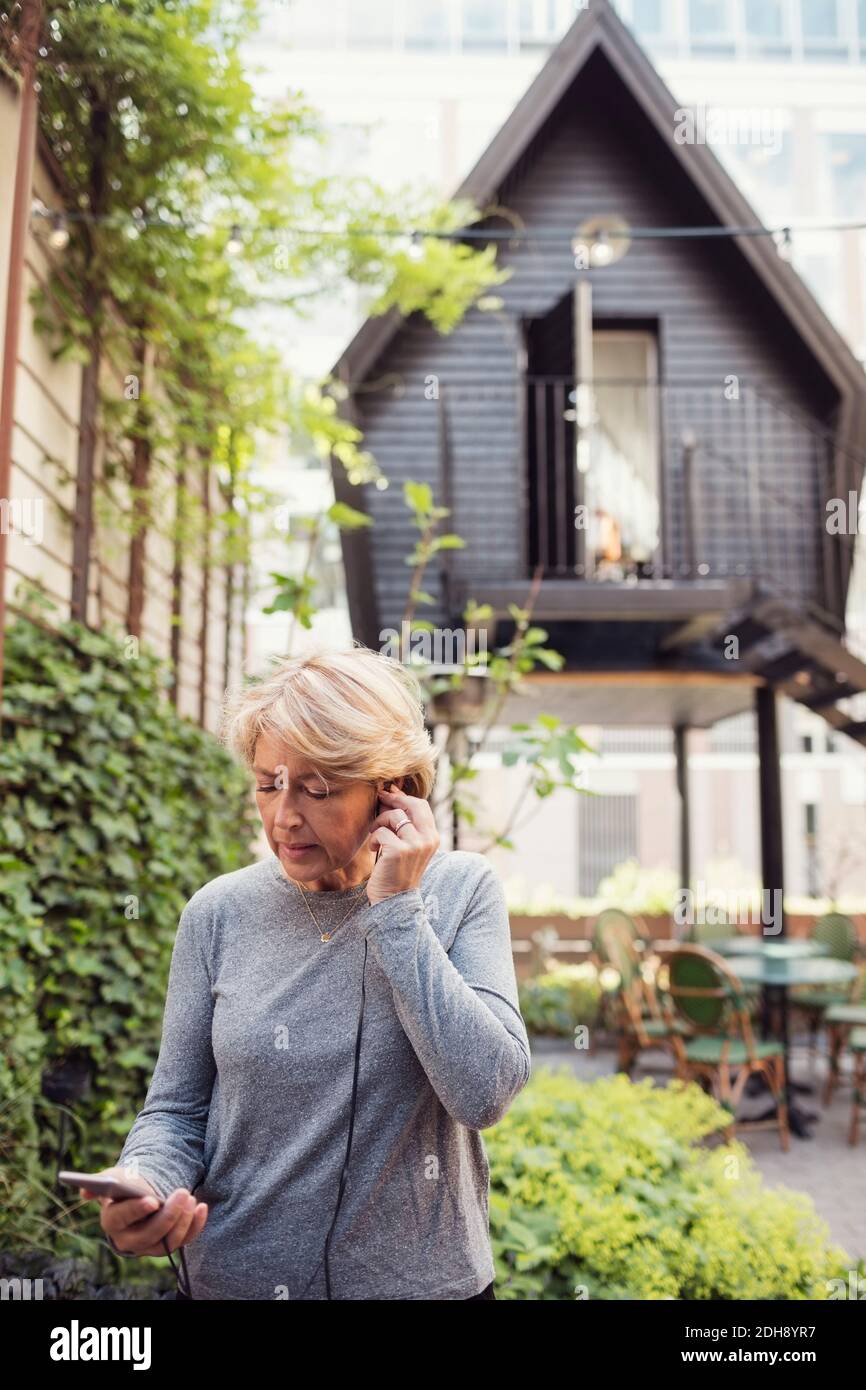 Donna d'affari matura che utilizza cuffie intrauricolari e smartphone in ufficio iarda Foto Stock