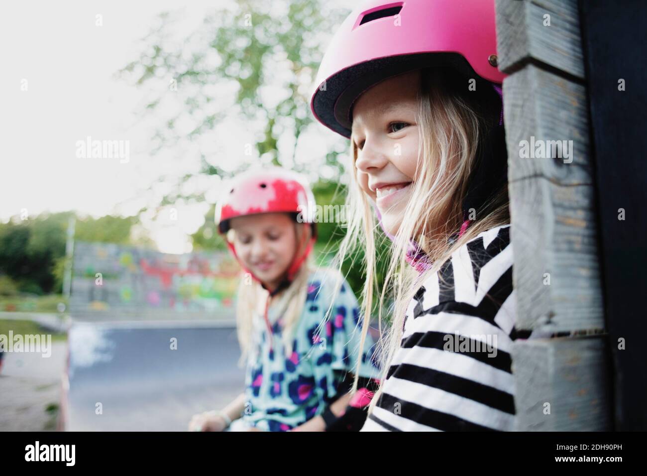 Ragazza sorridente che indossa il casco seduto con un amico sul bordo di rampa per skateboard Foto Stock