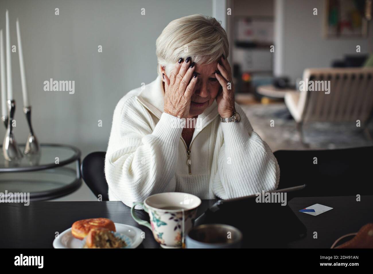 Donna anziana preoccupata con testa in mano guardando il digitale tablet mentre si siede al tavolo da pranzo a casa Foto Stock