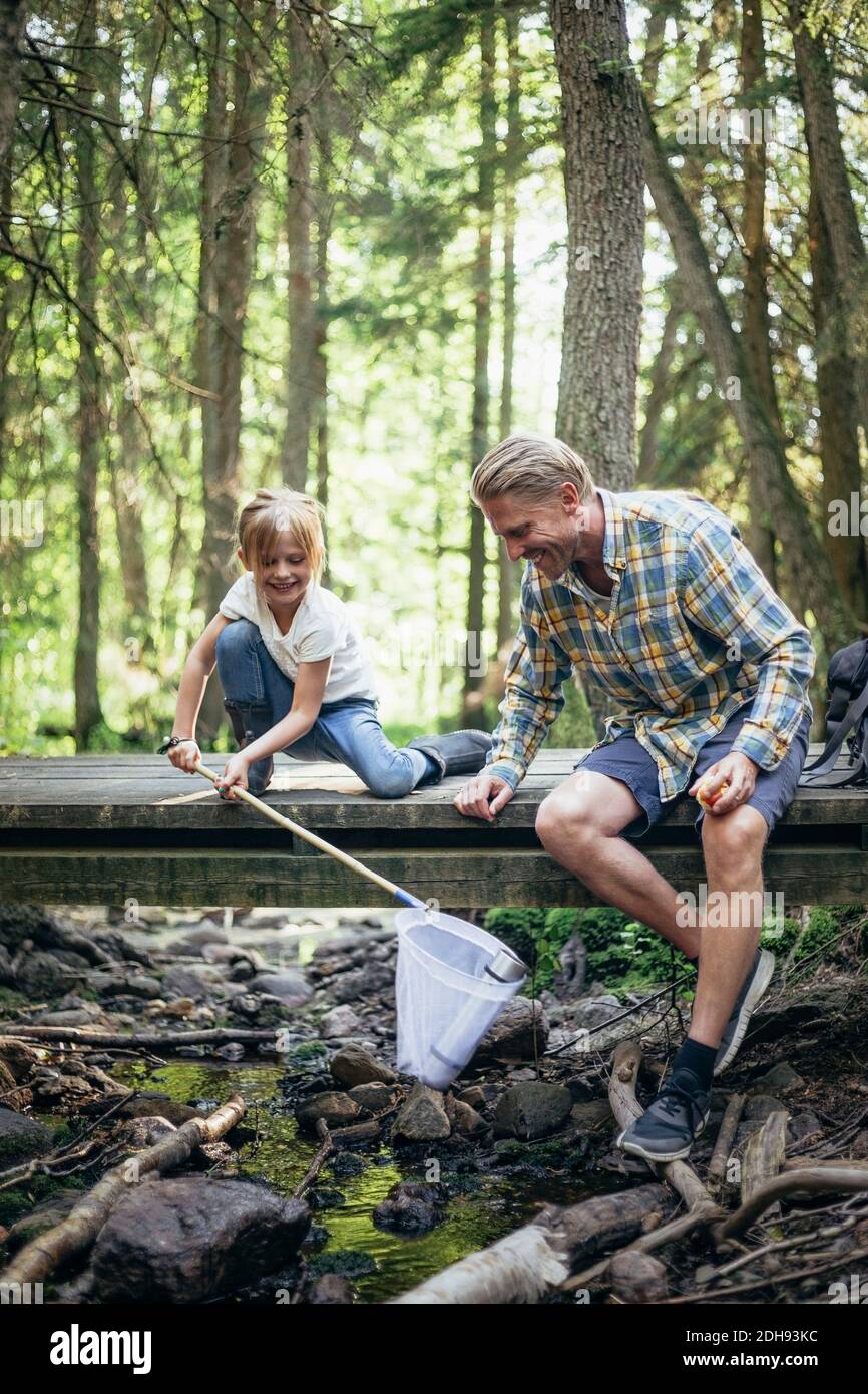 Figlia sorridente che tiene bottiglia in rete da pesca da padre in foresta Foto Stock